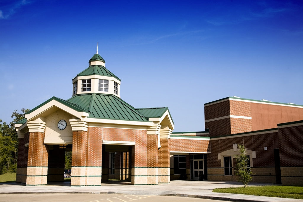 Front view of an elementary school building. Empty, no people. Clear blue sky. Red brick with green tin roof. Spire, clock.  Drop off area. Rotunda.